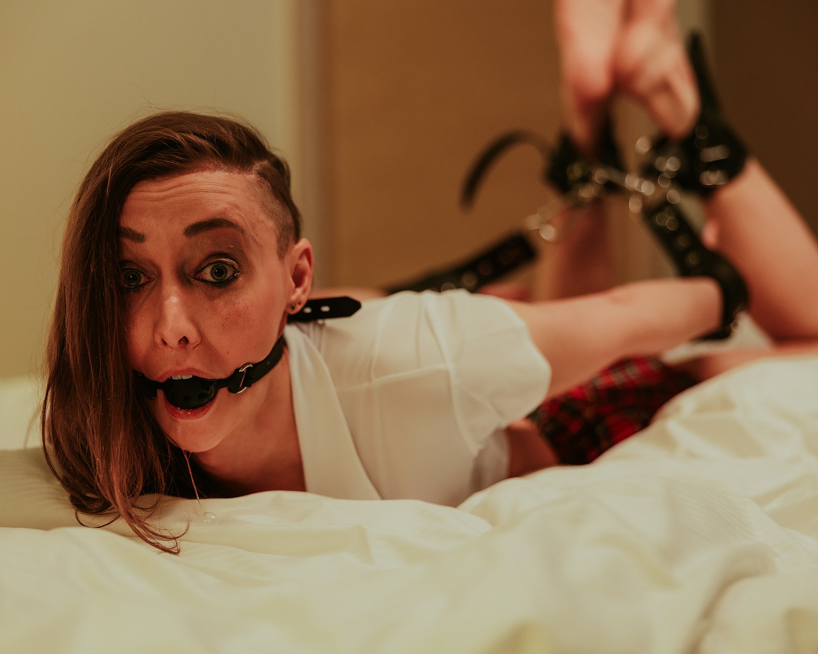 Jade koulutyttöasussa ja suukapulassa valkoisella hotellivuoteella alistumisen BDSM-kuvassa.